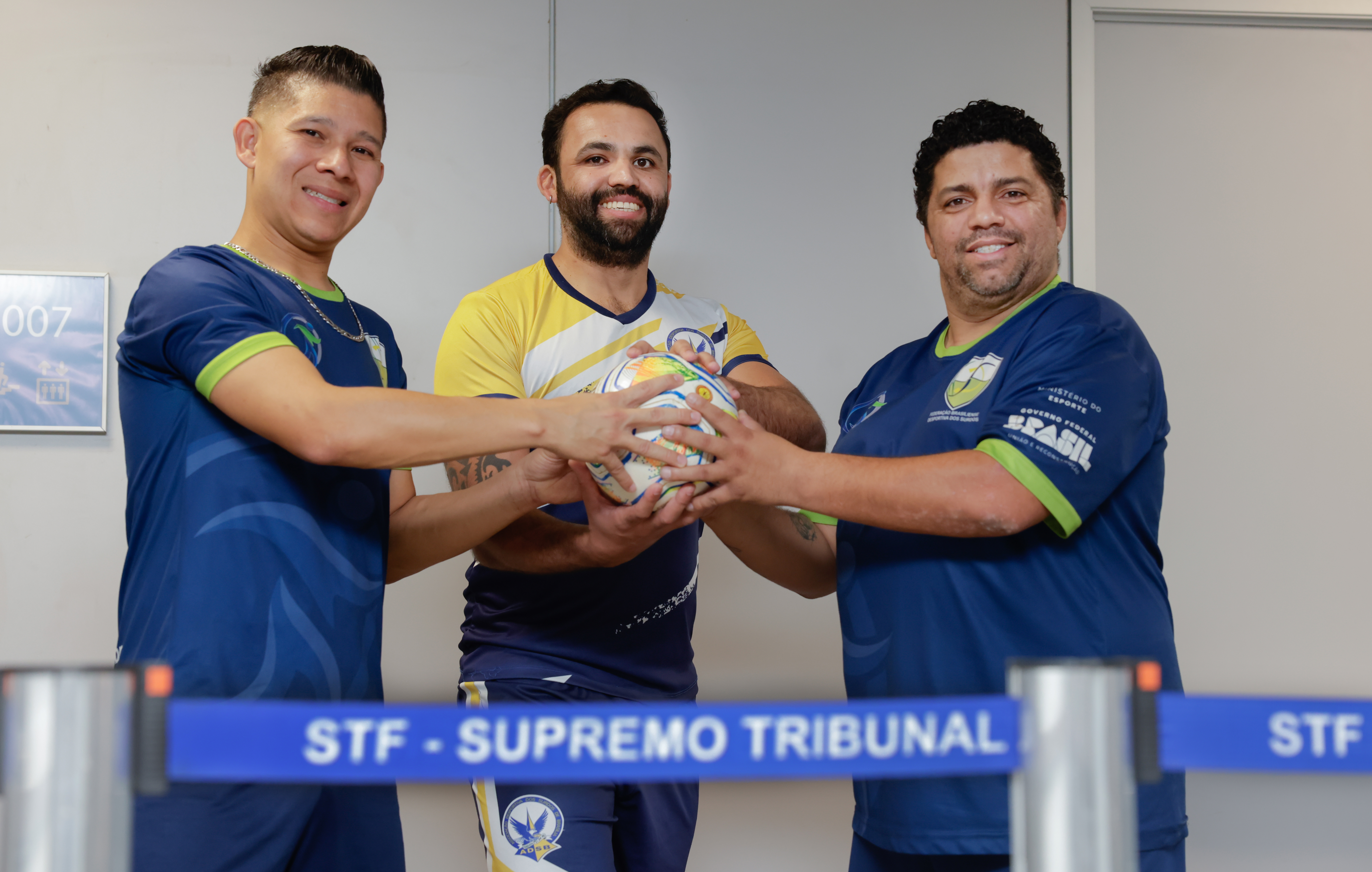 Foto colorida na horizontal mostra grupo de três homens que sorriem para a foto e seguram, juntos, uma bola de futsal, no meio da imagem. Da esquerda para direita, eles são Ederson Rocha, Joziel Nascimento e Aldo da Silva.