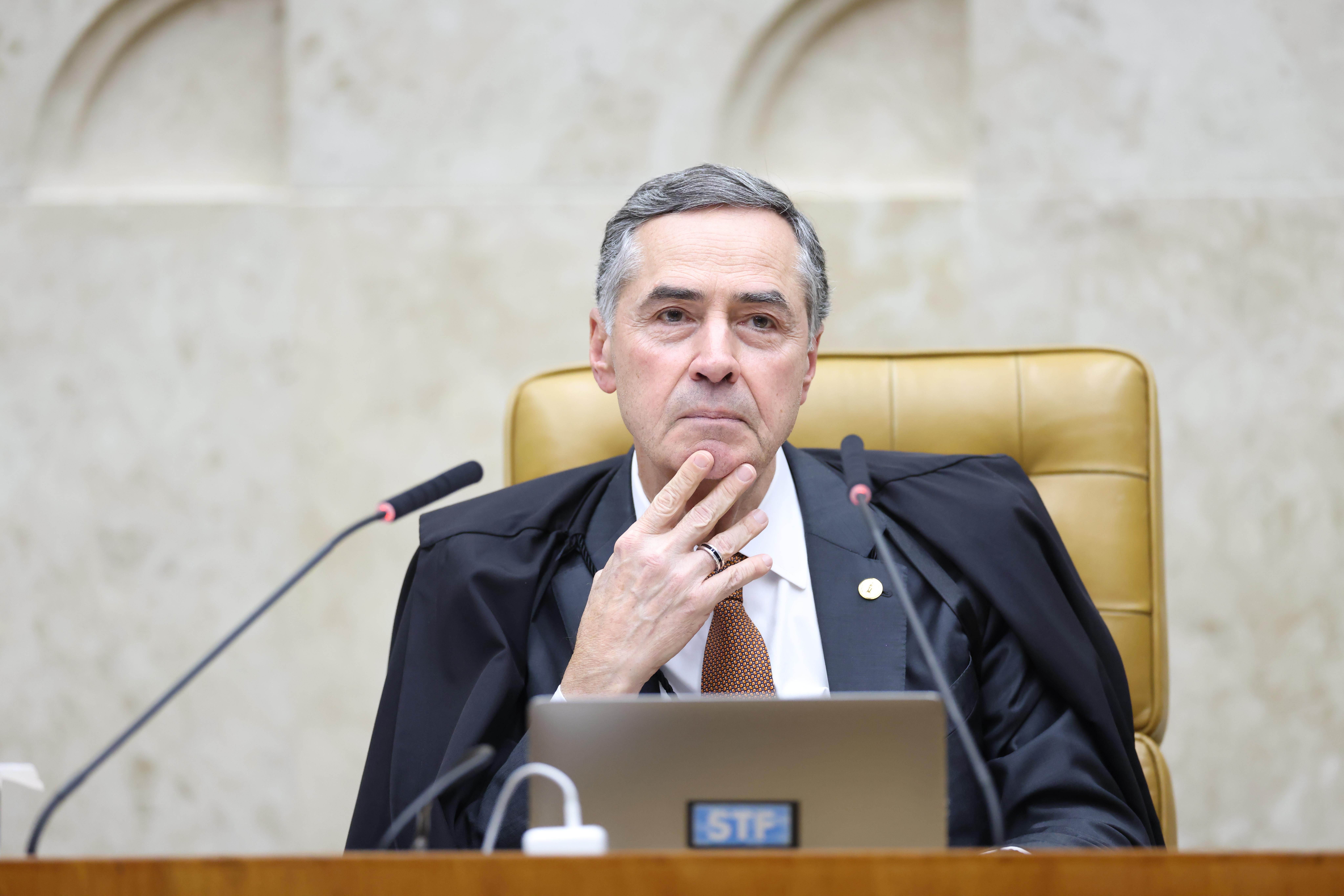 Fotografia colorida, na posição horizontal, do ministro Luís Roberto Barroso sentado na cadeira da Presidência do STF no Plenário. Ele está com a mão direita aberta tocando no queixo.