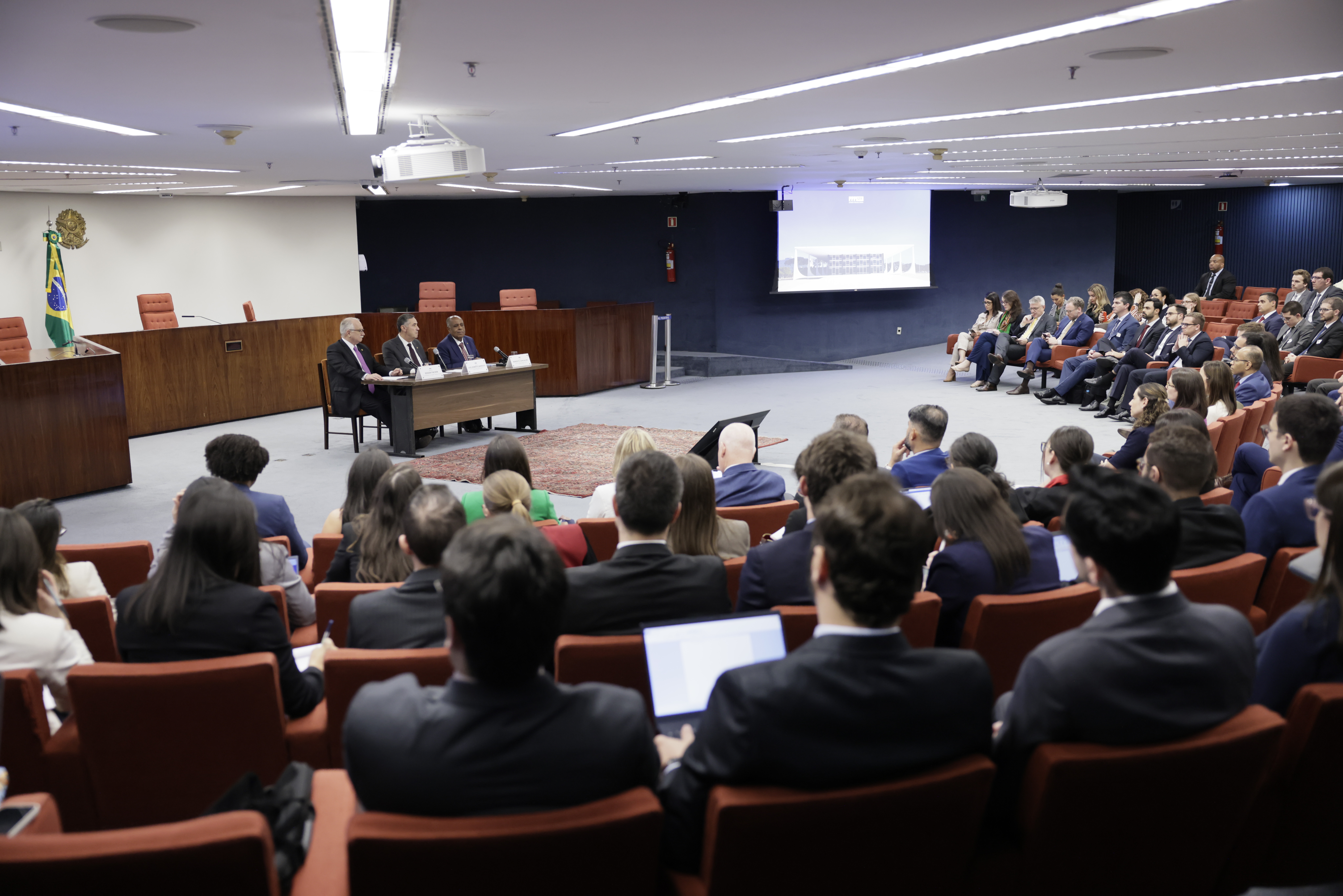 Foto colorida na horizontal da sala cheia dos novos magistrados sentados em poltronas vermelhas. na frente deles, numa mesa, estão o presidente do STF, ministro Luís Roberto Barroso, o vice-presidente, ministro Fachin, e o ministro Benedito Gonçalves, do Superior Tribunal de Justiça (STJ).