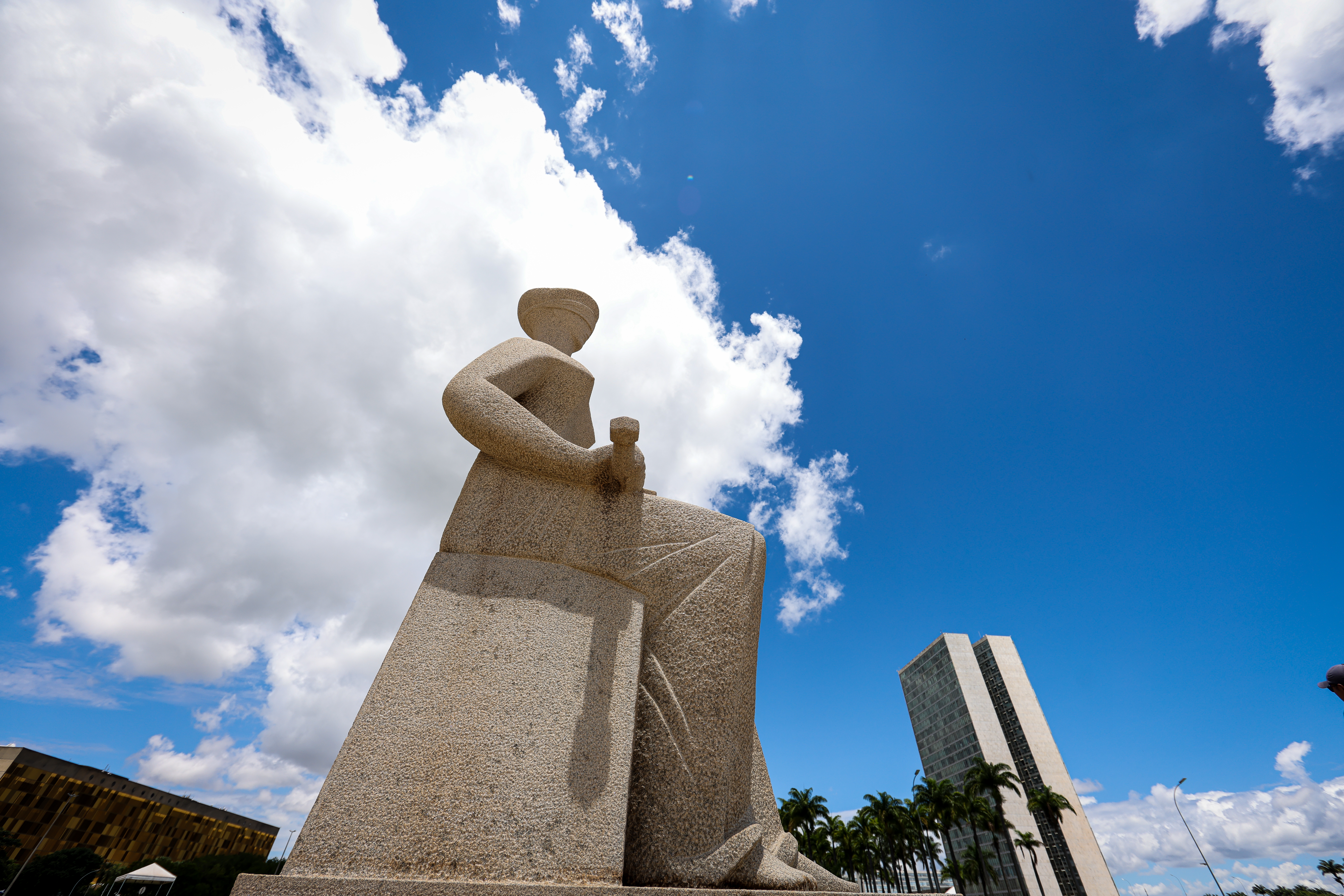 Foto em formato paisagem da estatua da Justiça de lado. A esquerda o prédio da Câmara dos Deputados e a direita o prédio do Congresso Nacional com palmeiras e o fundo céu azul